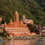 Scenic view of the riverside temple complex in Rishikesh with the Ganges River in the foreground, surrounded by lush green hills and peaceful Himalayan landscapes.