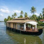 Traditional Kerala houseboat cruising through the peaceful backwaters, surrounded by coconut palms, calm canals, and lush tropical scenery on a sunny day.