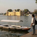 Traveler standing beside a traditional boat on the shores of Lake Pichola, admiring the iconic Lake Palace in Udaipur during a peaceful morning by the waterfront.