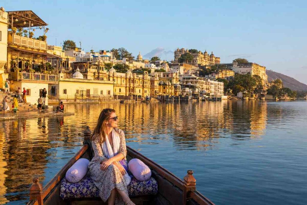 Tourist enjoying a traditional boat ride on Lake Pichola at golden hour, with Udaipur's historic waterfront, City Palace, and heritage buildings reflecting across the calm water.