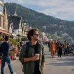 Foreign tourist enjoying a walk along Shimla's Mall Road with a coffee in hand, surrounded by colonial architecture, mountain views, cafés, and lively crowds on a sunny day.