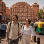International travelers exploring Jaipur's Hawa Mahal while walking through a vibrant market street surrounded by local shops, auto rickshaws, and Rajasthan's iconic Pink City architecture.