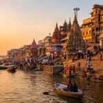 Sunrise over Varanasi's ghats with boats on the Ganges River, historic temples, and pilgrims gathering along the riverfront at dawn.